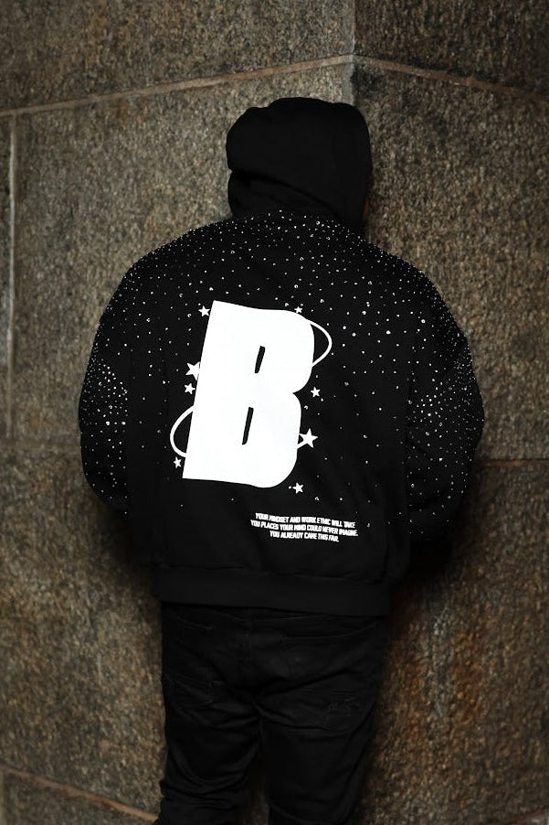 Person wearing a black hoodie with a large white letter 'B' on the back, standing against a textured wall.